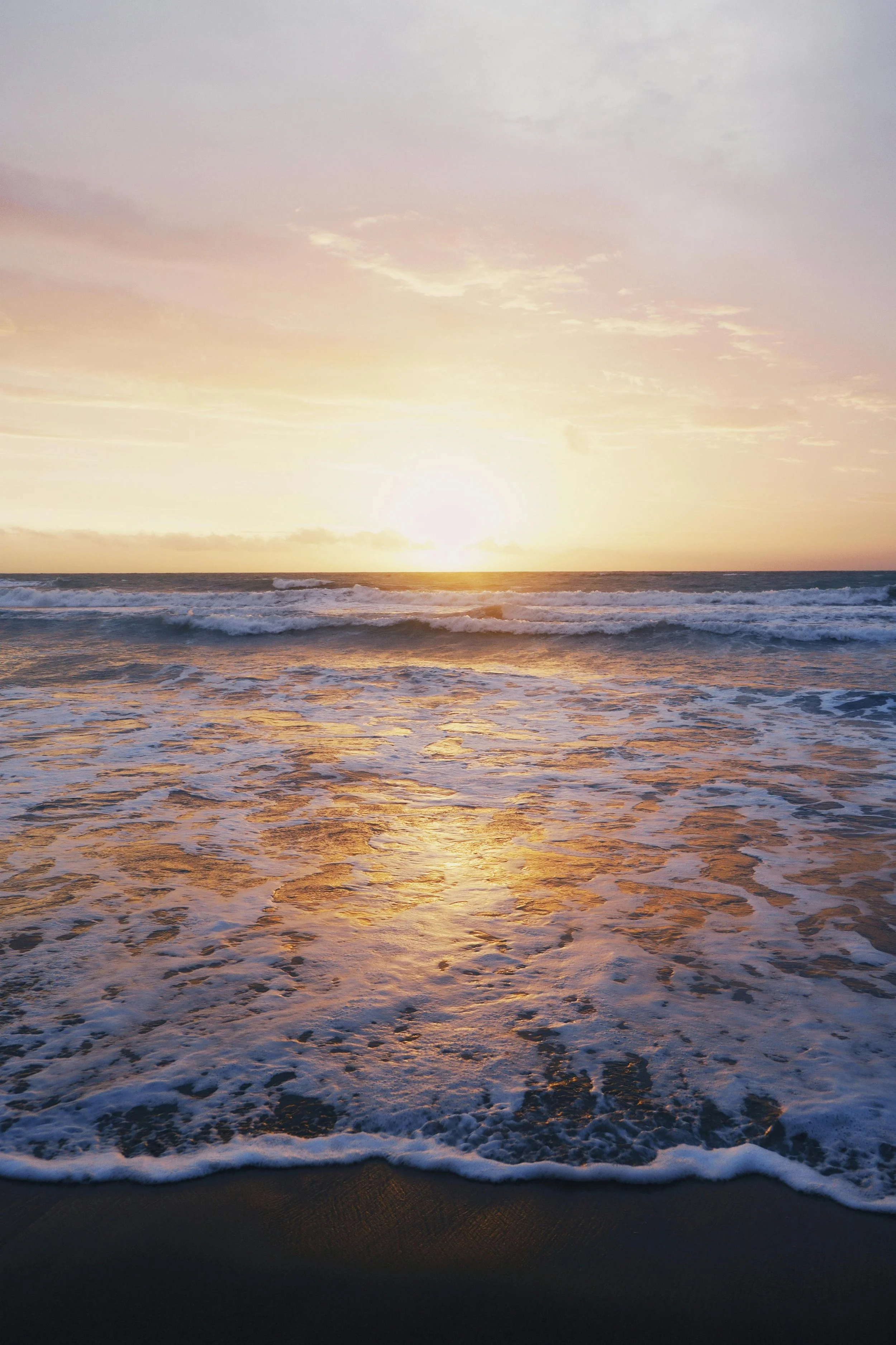 A sunset over the ocean with waves crashing onto the shore, reflecting the warm hues of the sky.