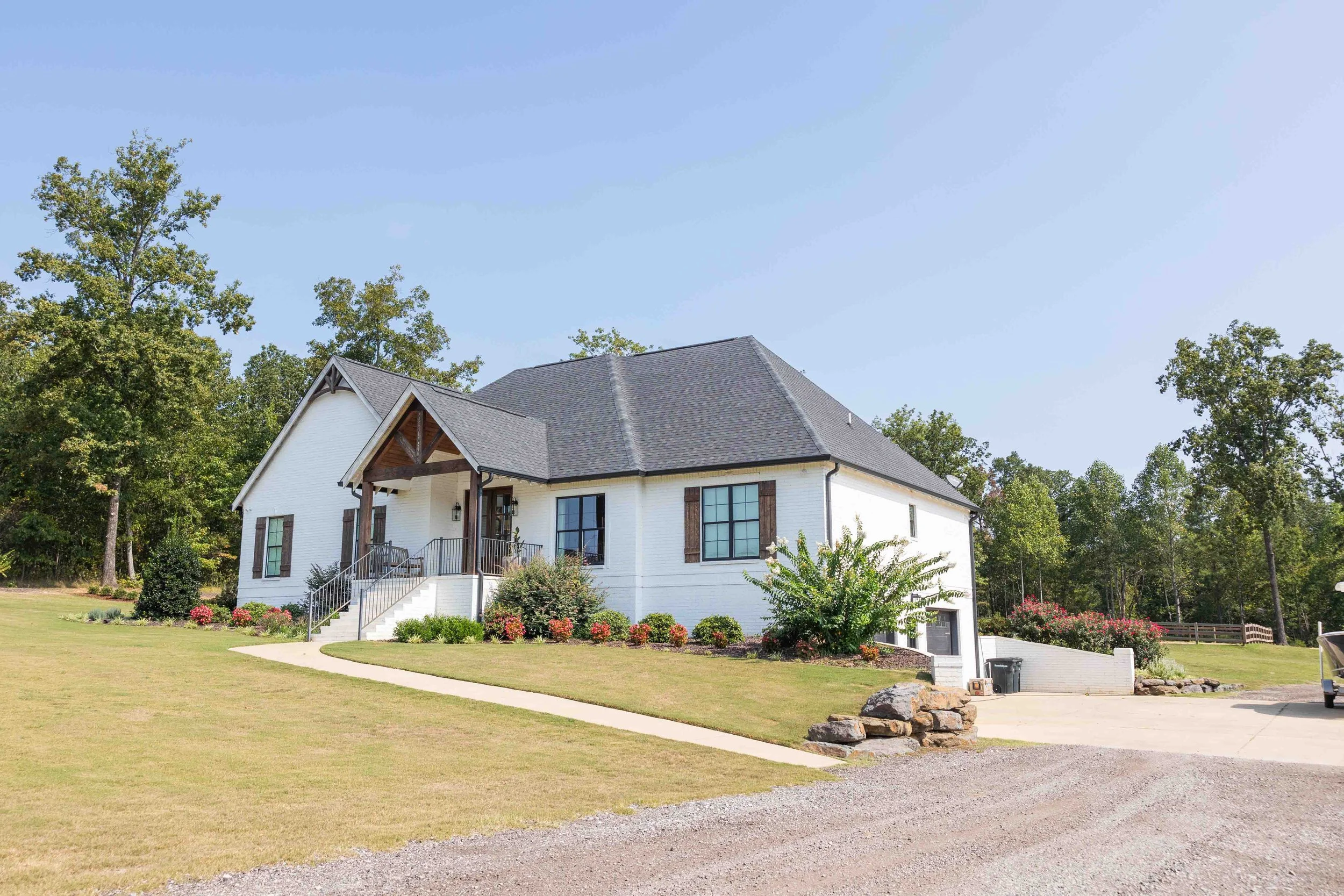 A white two-story house with a dark roof and front porch, surrounded by a manicured lawn and landscaped garden with bushes and flowers, set against a backdrop of trees and a clear blue sky.