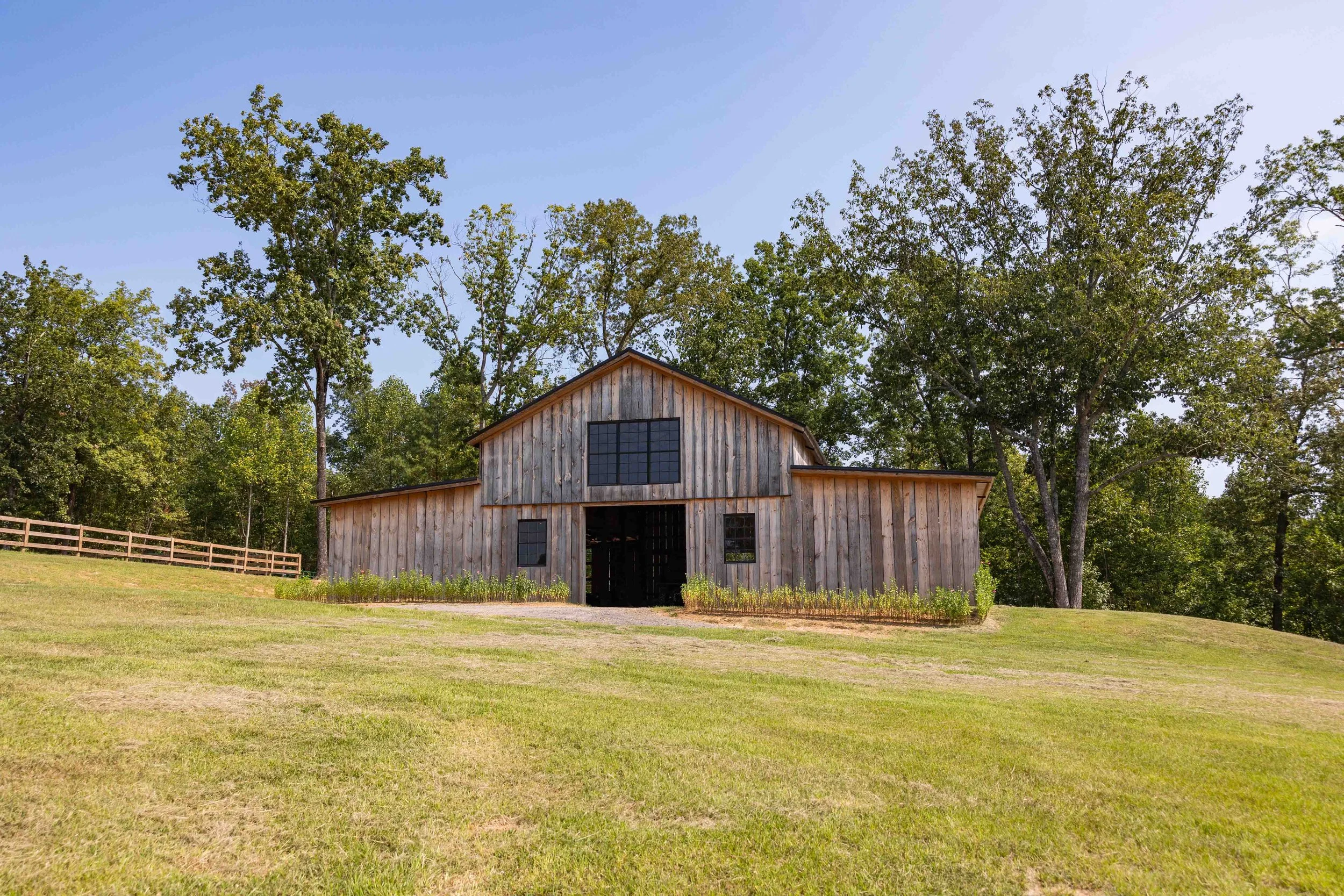 A rustic wooden barn in a grassy field with trees in the background under a clear blue sky.