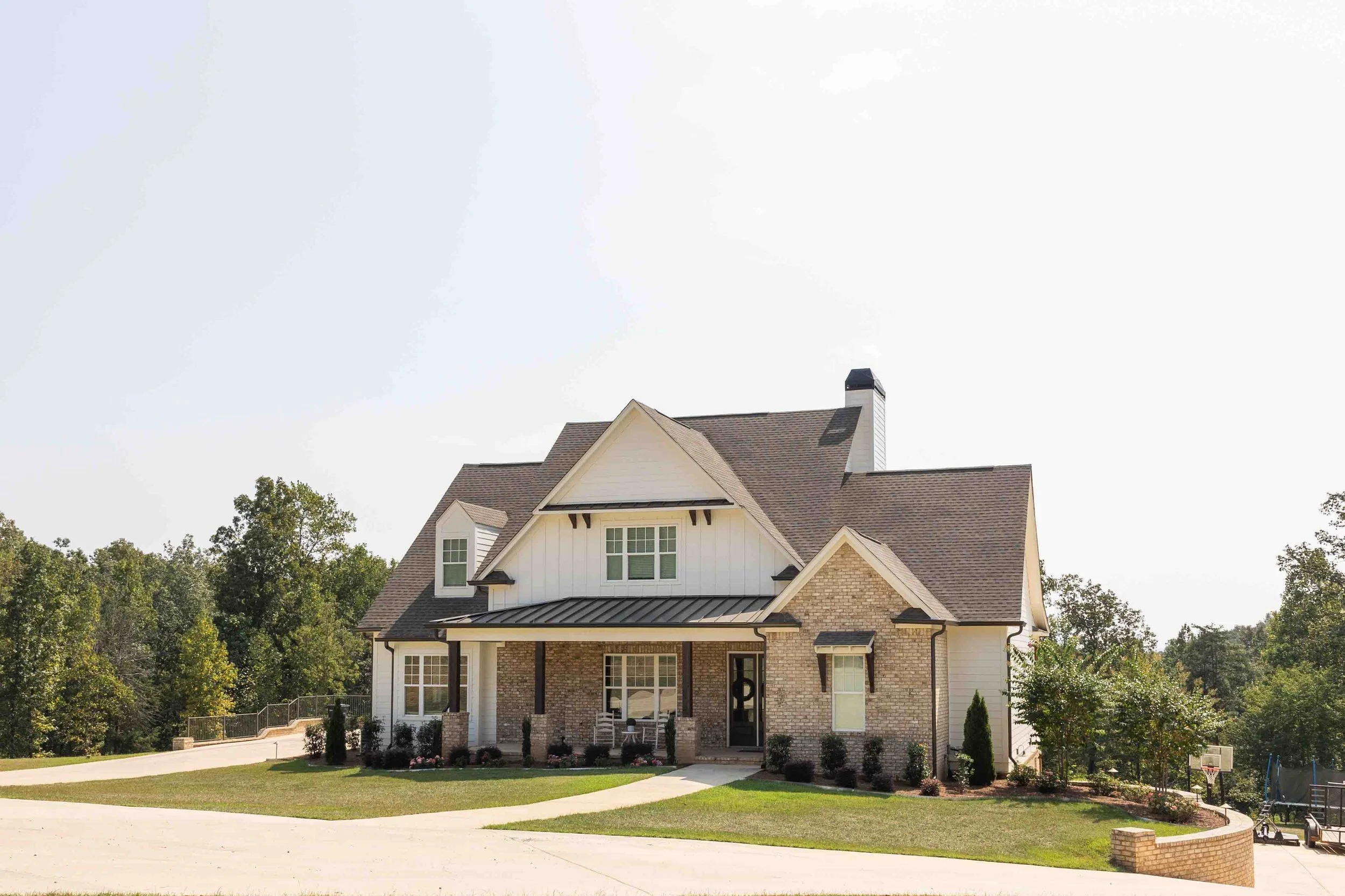 Beautiful two-story house with a blend of brick and white siding, front porch with seating, surrounded by well-maintained lawn and trees in the background.