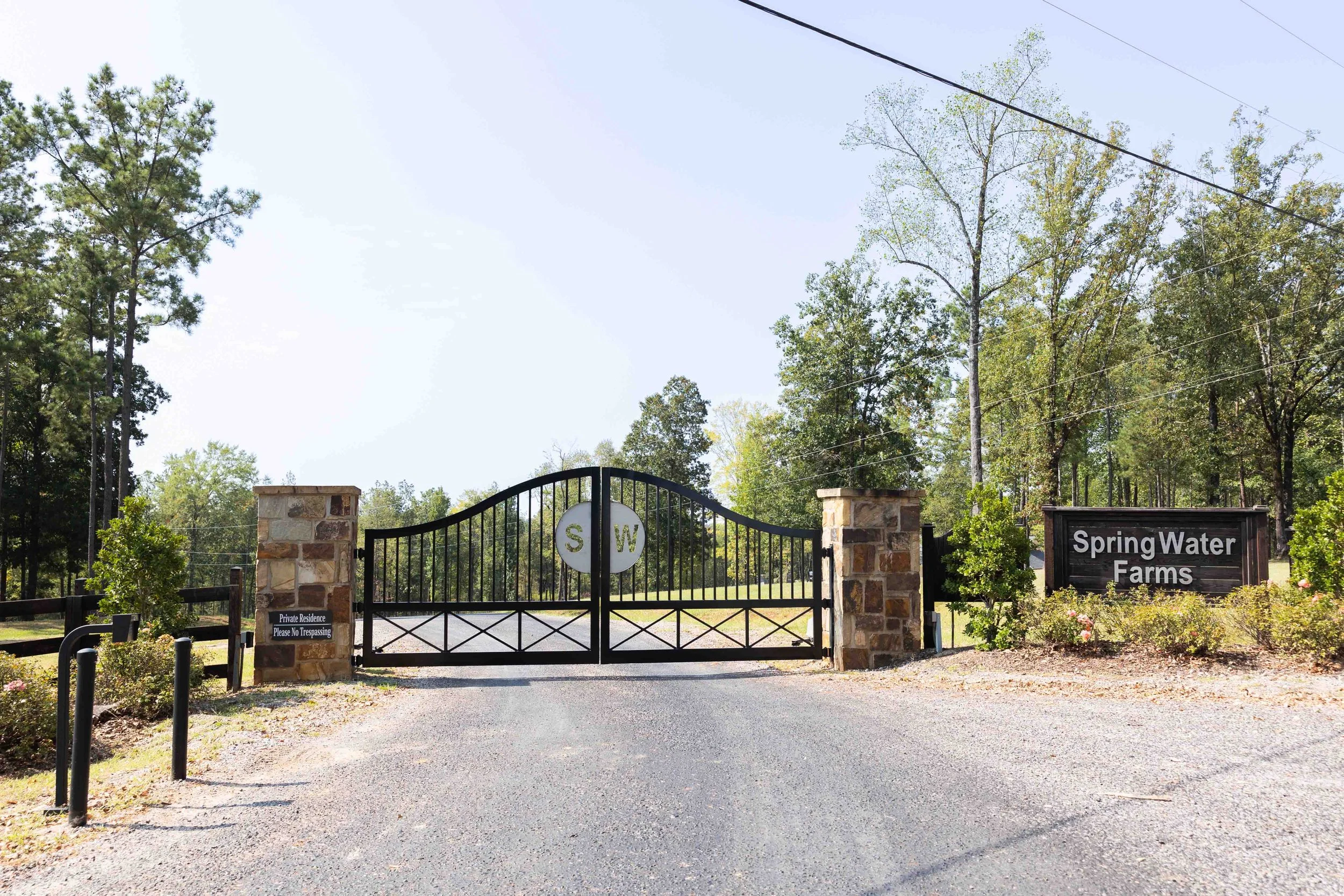 Entrance gate to Spring Water Farms with stone pillars, black metal gate with 'S' and 'W' initials, and signs indicating private residence and no trespassing, surrounded by trees and greenery.