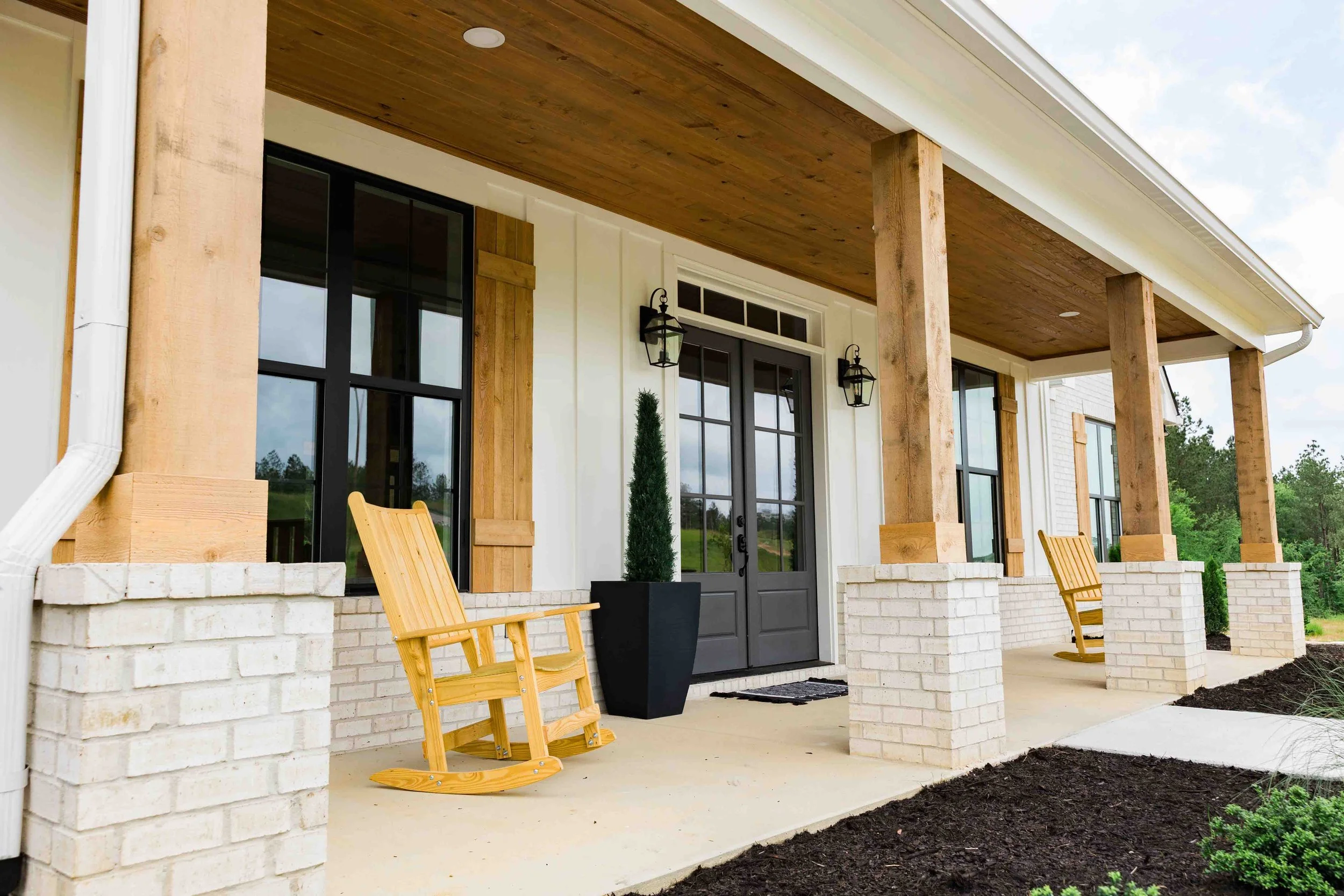 Front porch of a house with white brick columns, wooden posts, and a wooden ceiling. There are two wooden rocking chairs, a tall potted shrub, and black lantern-style wall lights. The house has black-framed glass doors and windows, with a grassy land