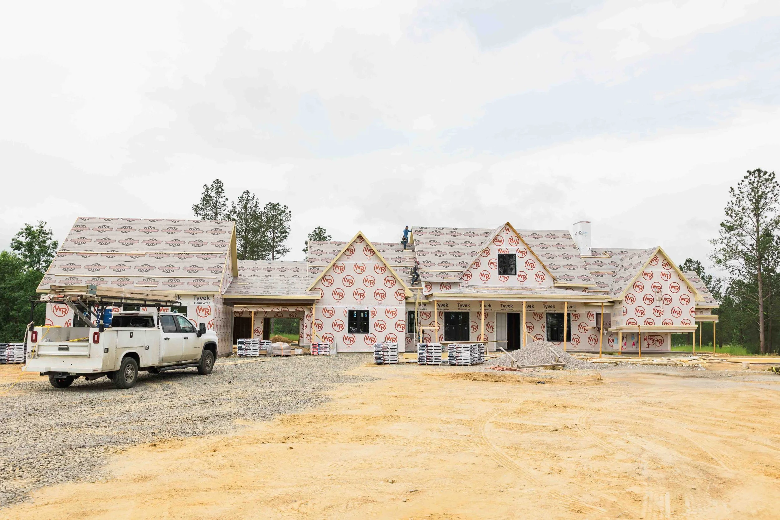 A large house under construction with the exterior still covered in specialized building wrap, and construction workers on the roof. There are construction materials and a white pickup truck parked in front on a dirt and gravel lot, with trees in the