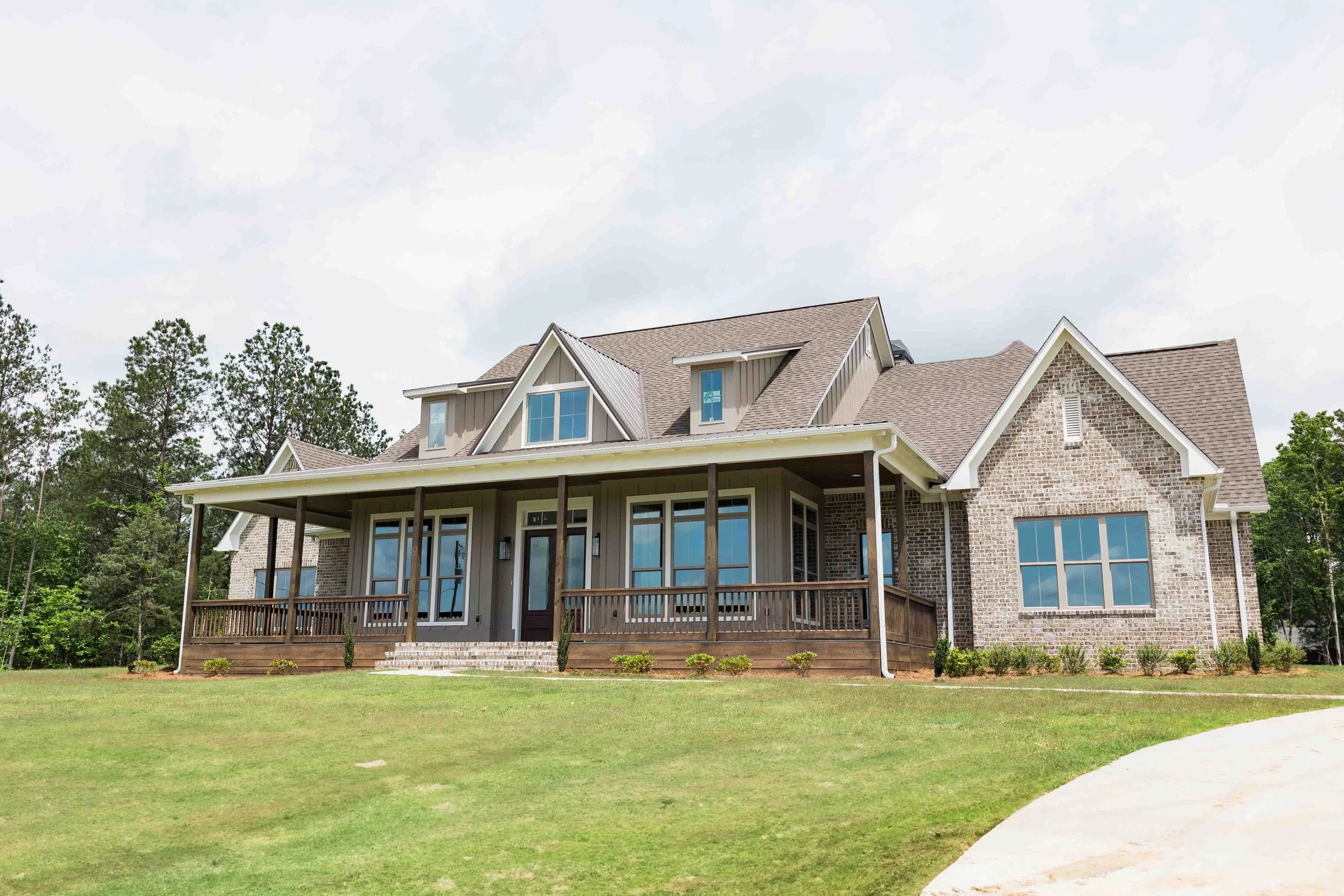 A large two-story house with a brick and gray siding exterior, a covered front porch, and multiple gabled roofs, surrounded by a green lawn and trees under a cloudy sky.