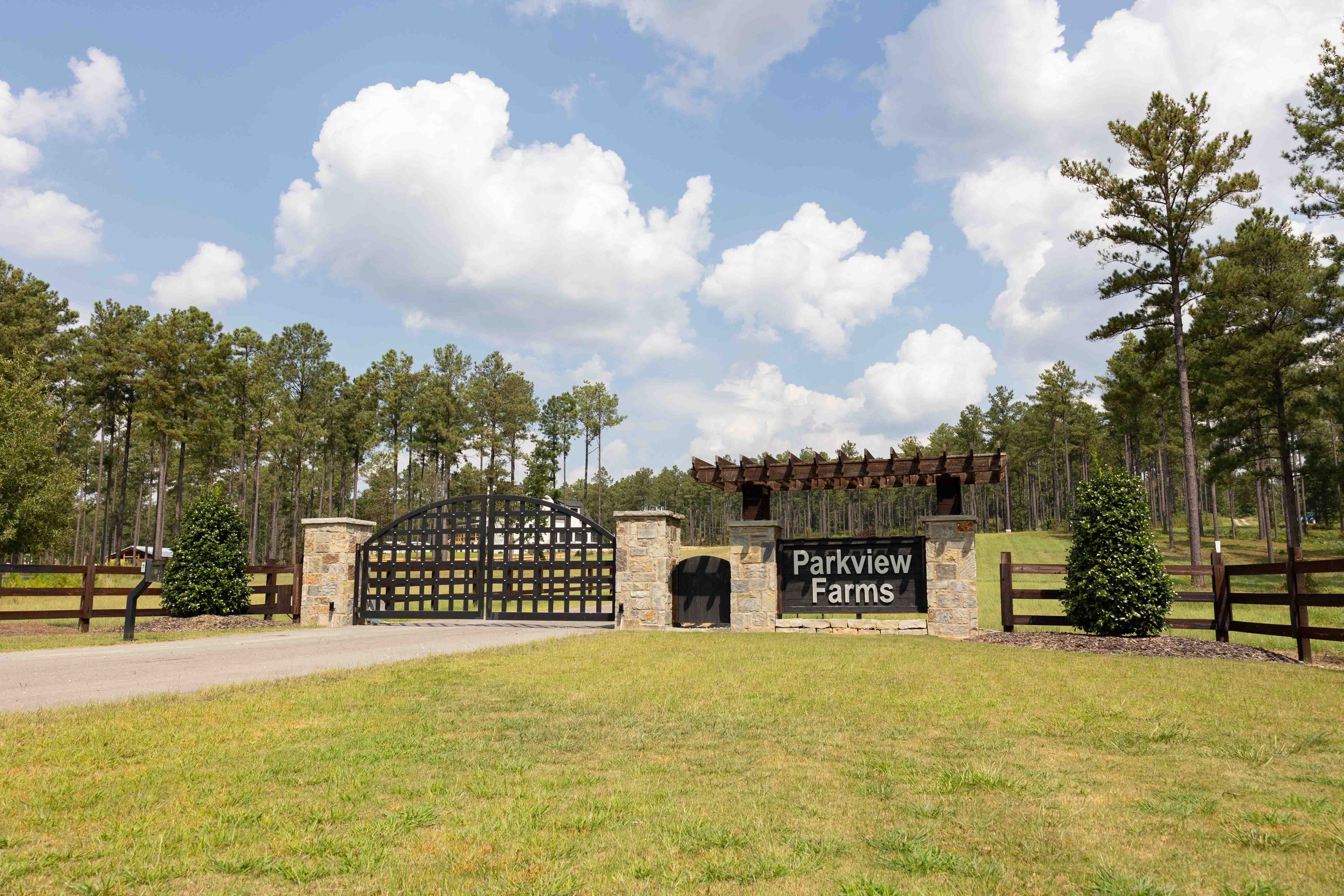 Entrance gate to Parkview Farms, with a black metal gate, stone pillars, and a sign reading "Parkview Farms." There are trees and a grassy area, with a blue sky and scattered clouds in the background.