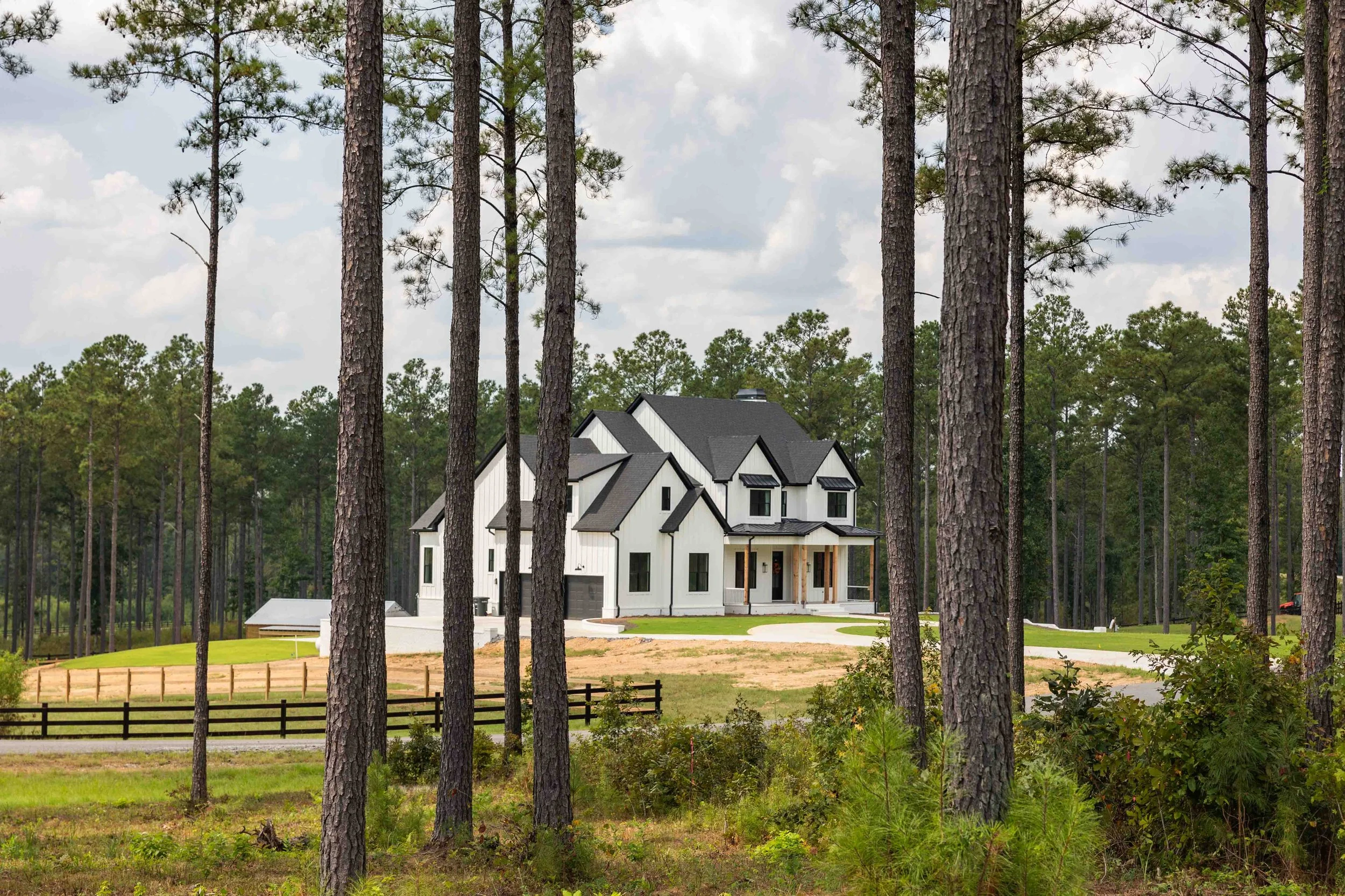 A large, modern white house with black roof accents situated among tall pine trees on a green lawn.