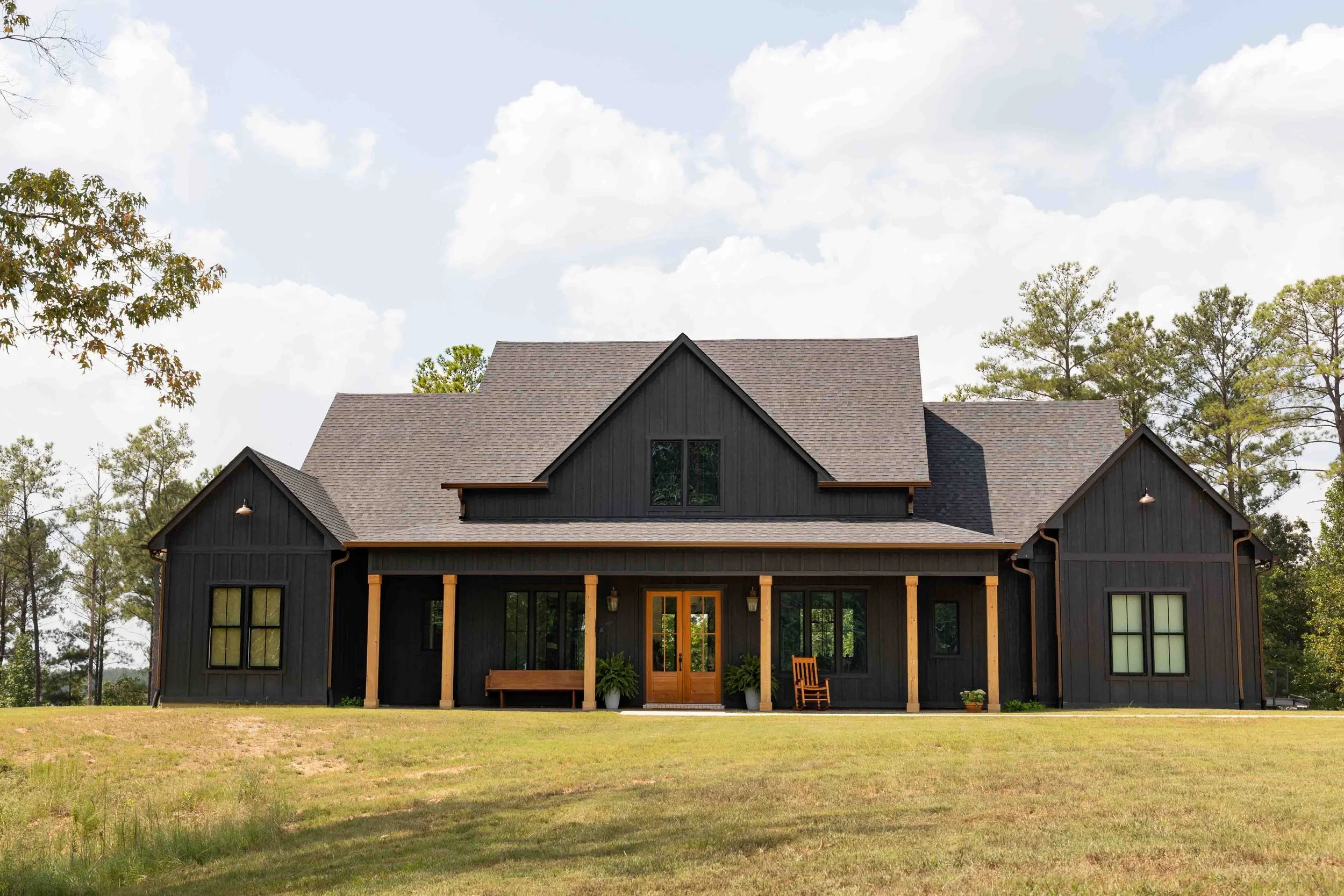 A black farmhouse-style home with wooden accents, a large front porch with columns, and surrounding trees.