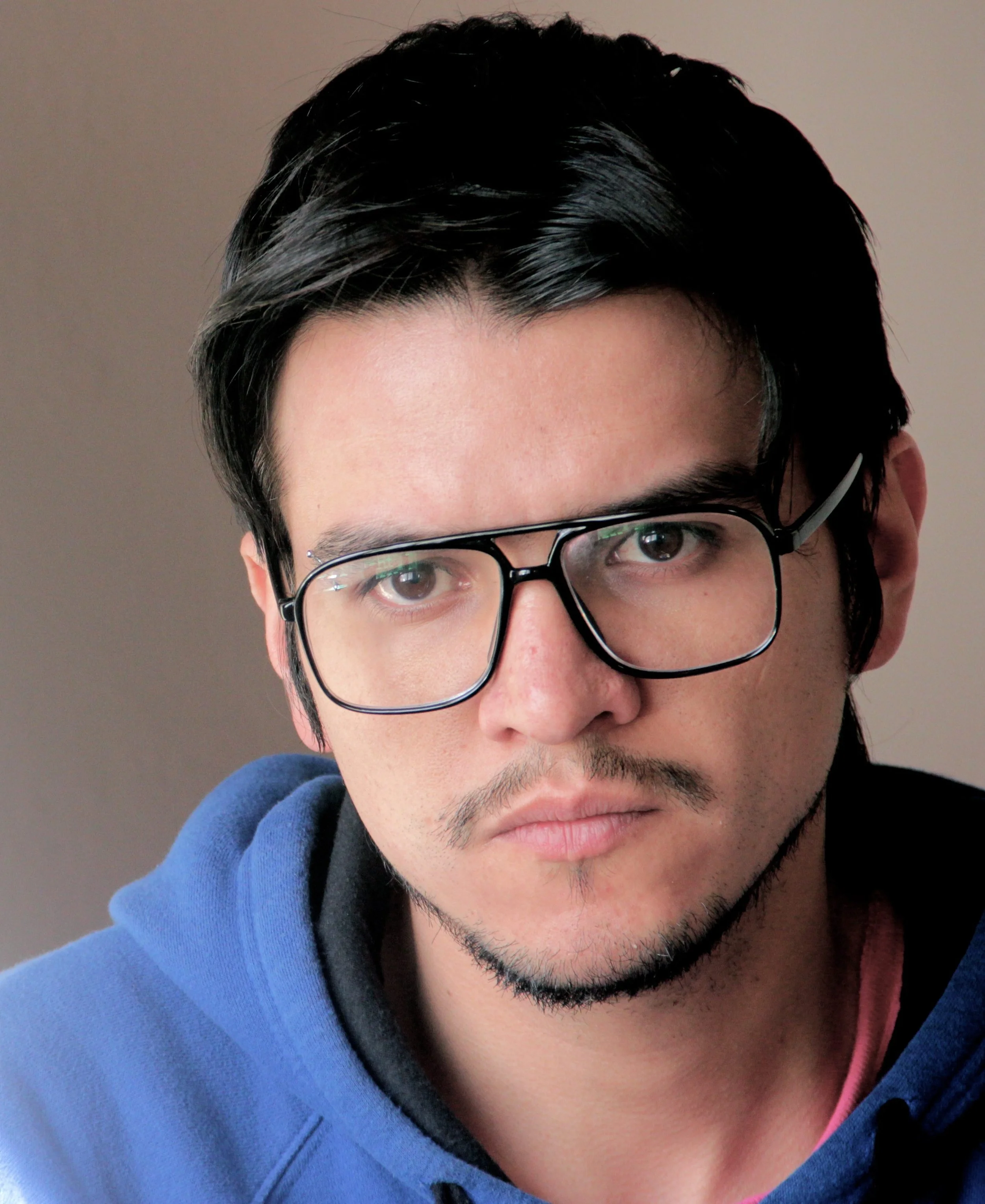 Gonzalo Escobar. Director of photography and editor of the film. A young man with black hair, wearing glasses, a faint mustache, and a goatee, dressed in a blue hoodie, looking directly at the camera with a neutral expression.