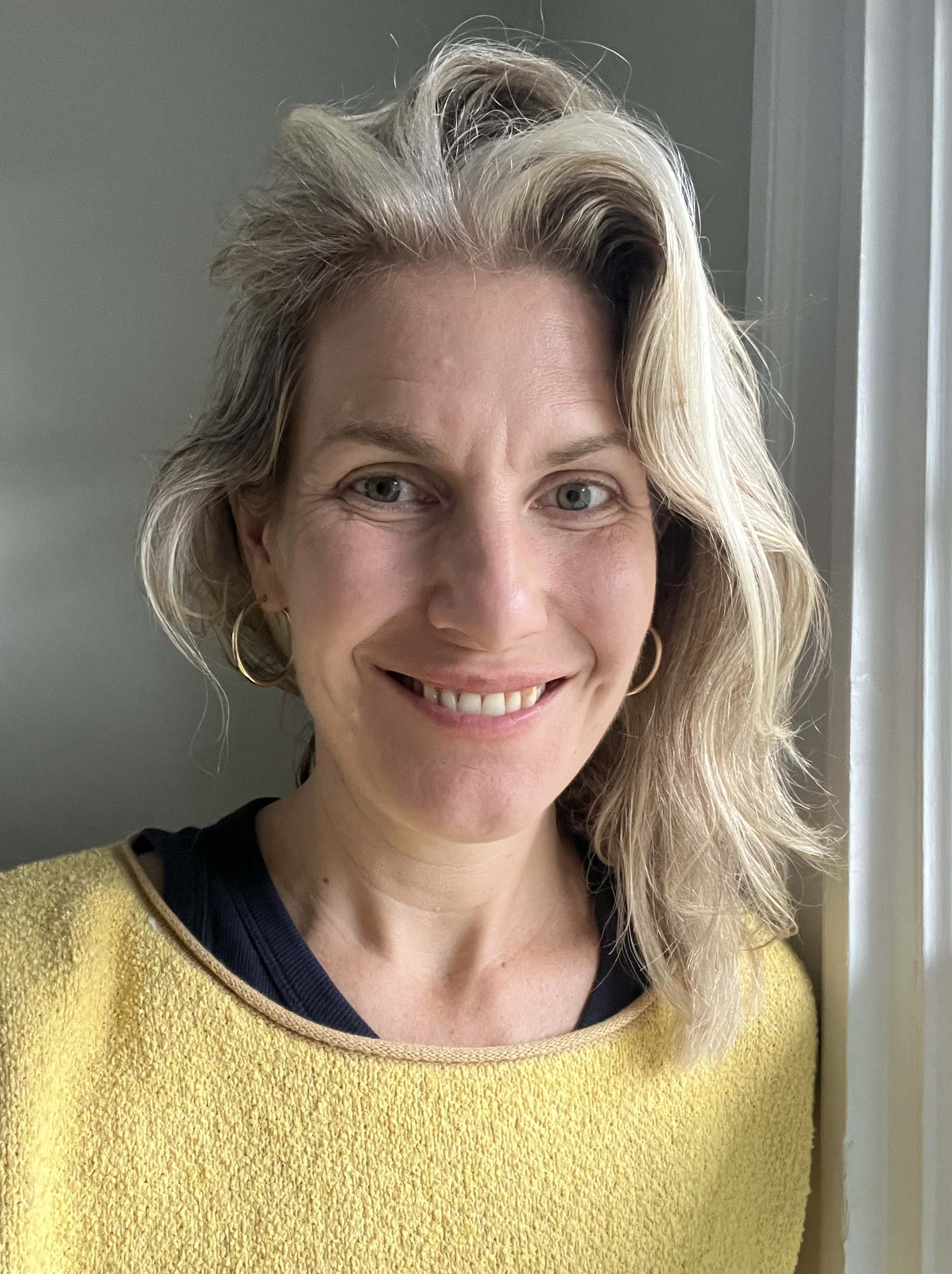 Brenda Ávila-Hanna. Executive producer of the film. A smiling woman with blonde hair, wearing hoop earrings and a yellow top, standing near a window.