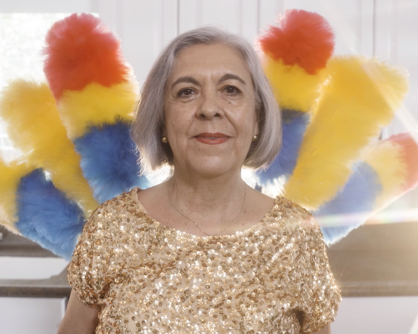 Beatriz Valencia. A woman smiling and wearing a sequined dress. In the background are several feather dusters in vibrant colors.