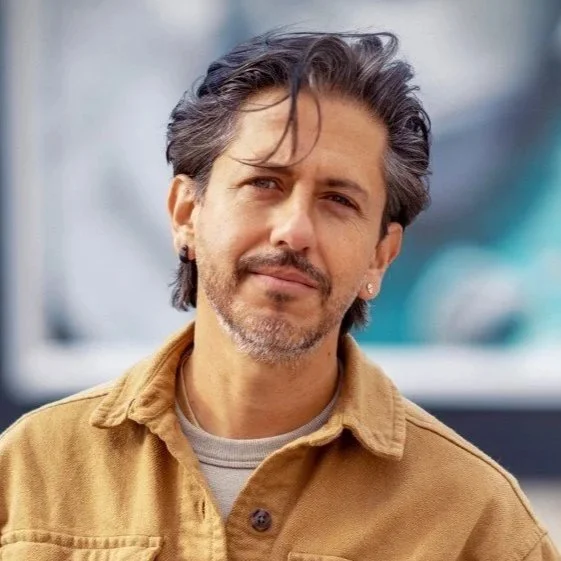 Juan Mejia. Executive producer of the film. A man with gray hair and a beard, wearing a beige jacket and a gray shirt, standing outdoors with a blurred background.