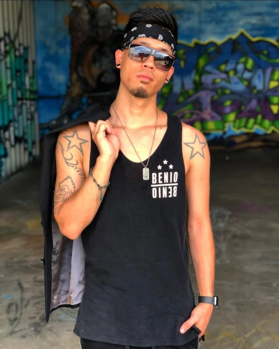A young man with tattoos on arms, wearing sunglasses, a black bandana, a sleeveless black shirt, a necklace, and a wristwatch, holding a jacket over his shoulder, standing in front of graffiti art.