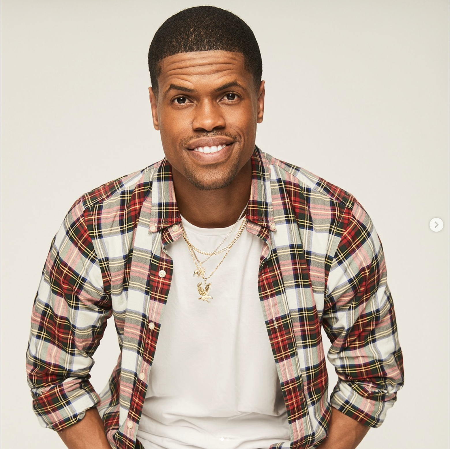 Young man with short dark hair, wearing a white T-shirt, layered with a red, black, yellow, and white plaid shirt, smiling, with necklaces, against a plain off-white background.