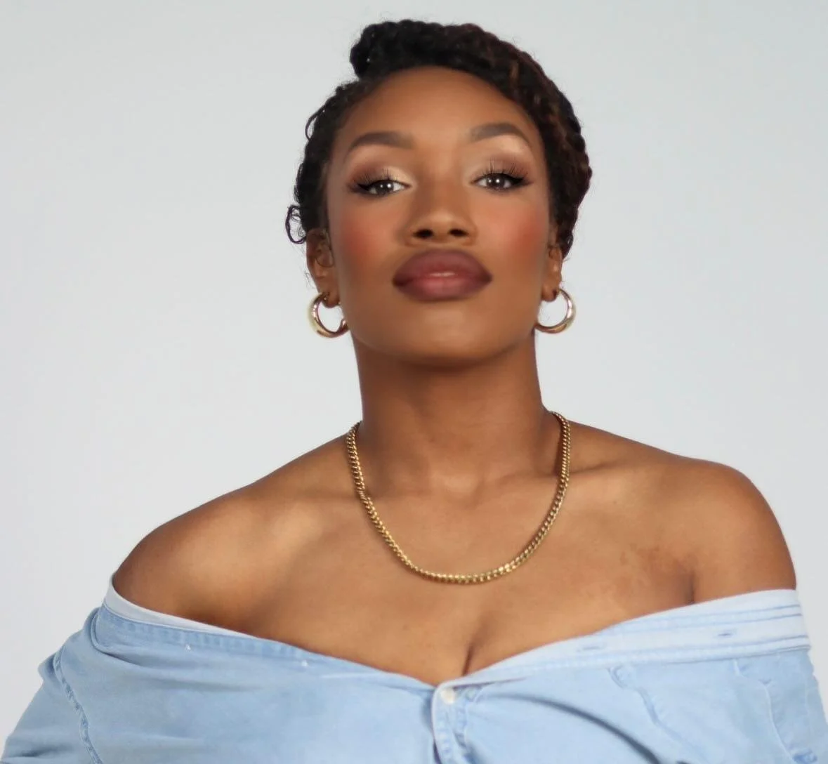 Portrait of a woman with short twisted hair, hoop earrings, gold chain necklace, wearing an off-the-shoulder light blue shirt against a plain light background.