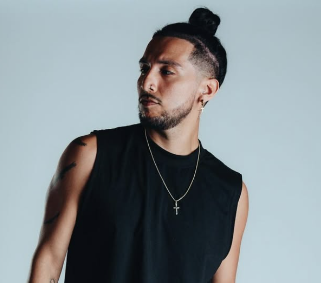 Man with long dark hair in a bun, wearing a sleeveless black shirt and a cross necklace, posing against a light blue background.