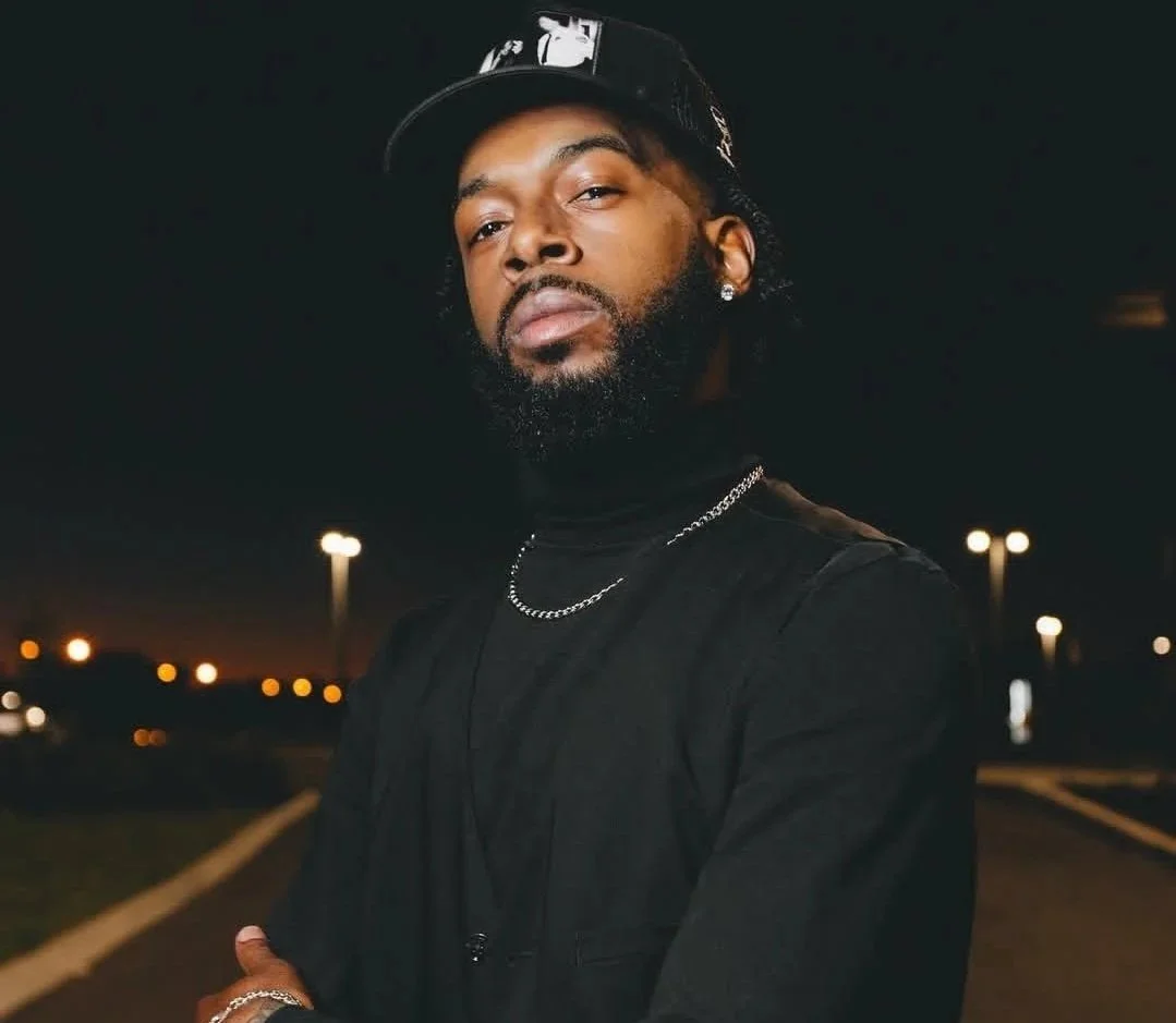A man with a beard and mustache wearing a black cap, black shirt, and silver jewelry standing outdoors at night with blurred streetlights in the background.