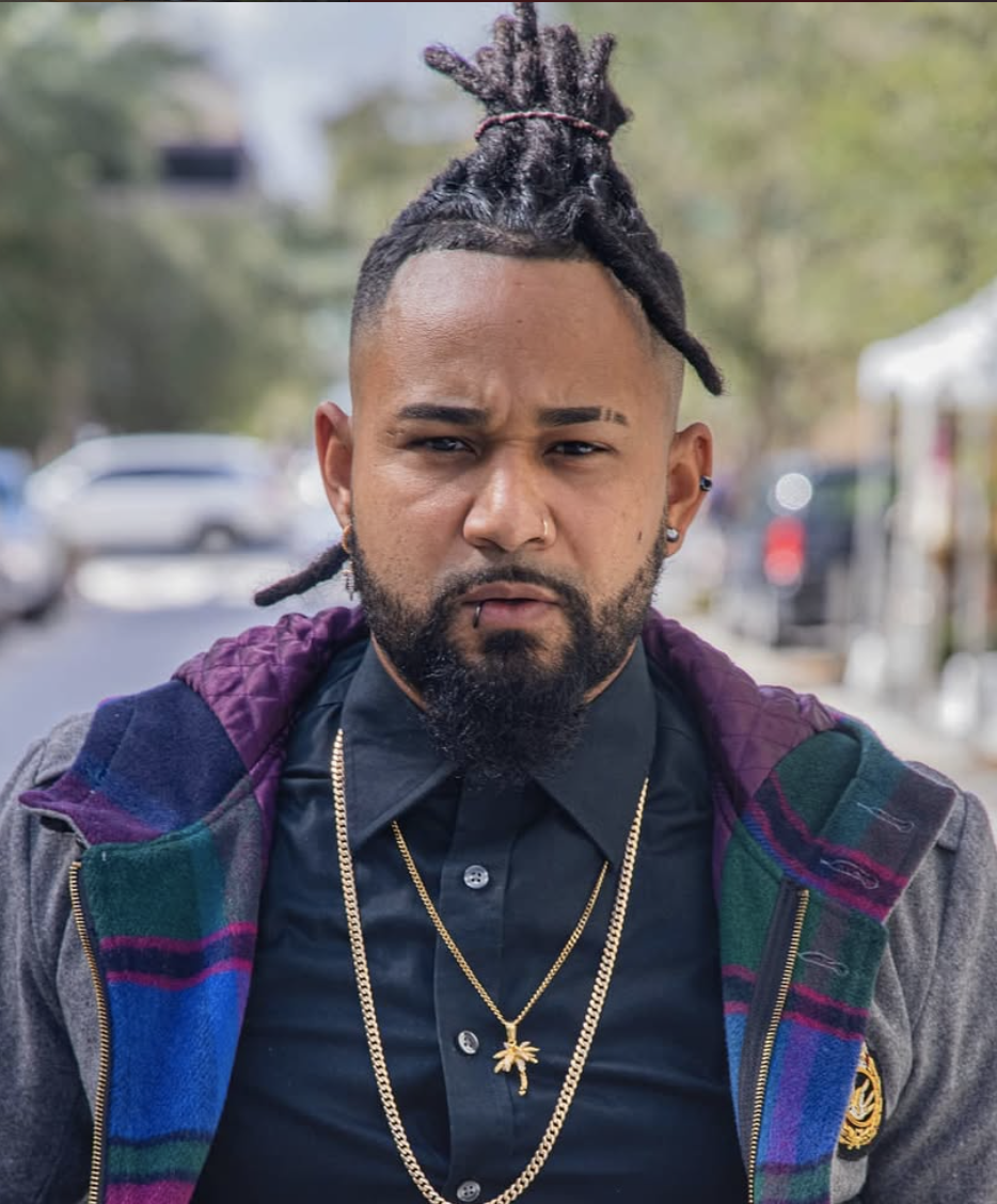 A man with dreadlocks tied up with a red hairband, beard, and piercings, wearing a black shirt and layered gold necklaces with a marijuana leaf pendant, outdoors on a street with cars and trees in the background.