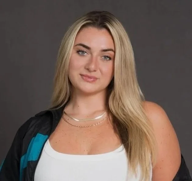 A young woman with long blonde hair, light skin, wearing a black jacket with blue accents over a white tank top, posing against a gray background.
