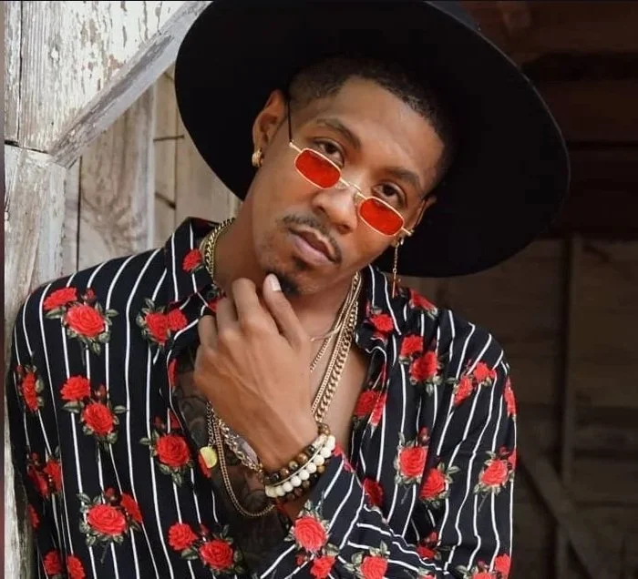 Man wearing a black hat, red sunglasses, and a black shirt with red roses and white stripes, standing in front of wooden wall.