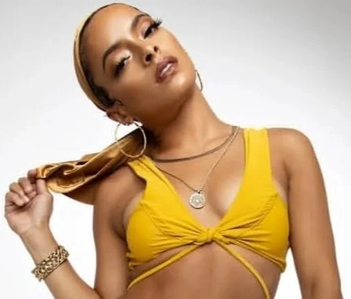 A woman with long hair tied with a scarf, wearing a yellow crop top, gold jewelry, and posing with her head tilted.