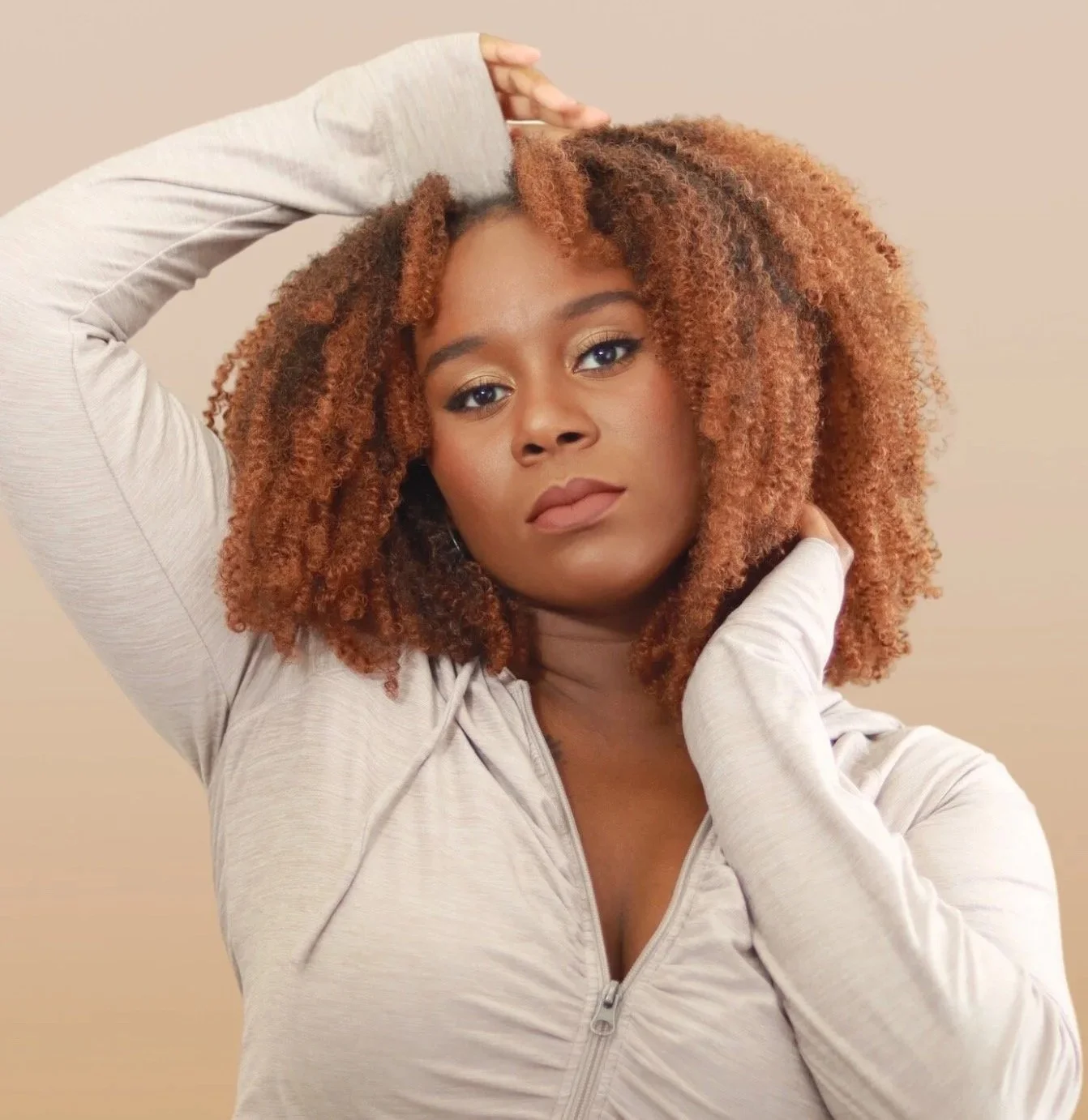 Portrait of a woman with curly, reddish hair wearing a light-colored zippered jacket, looking at the camera with a neutral expression.