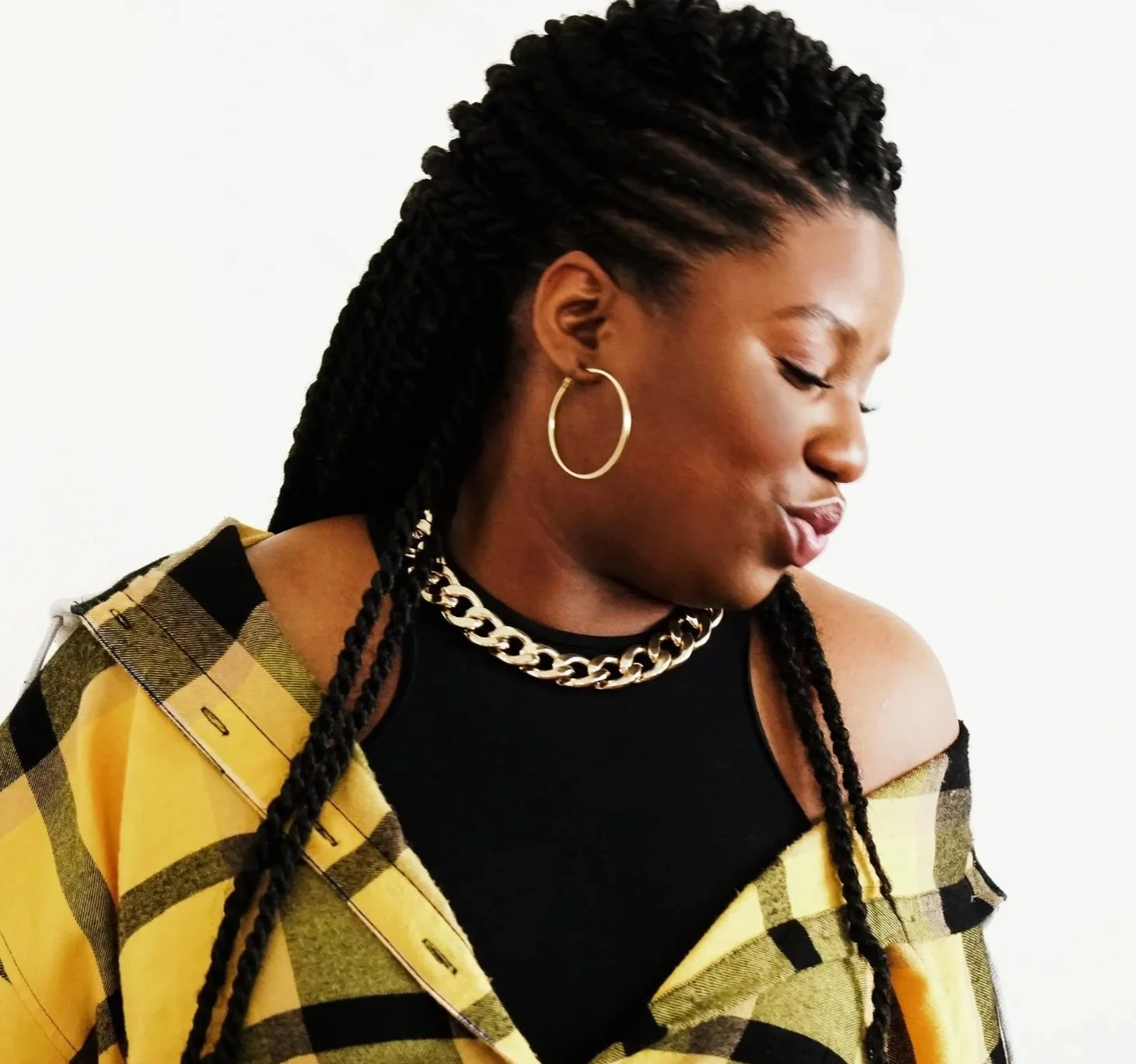 A young woman with braided hair, wearing gold hoop earrings, a chunky gold chain, and a black top with a yellow and black plaid shirt off the shoulder.