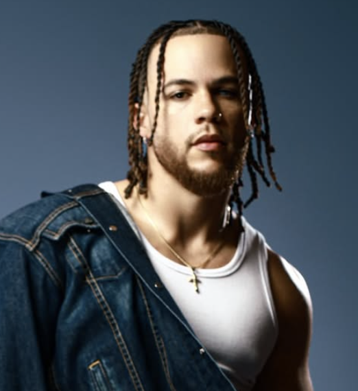 Young man with braids, beard, and a nose ring wearing a white tank top and denim jacket against a blue background.