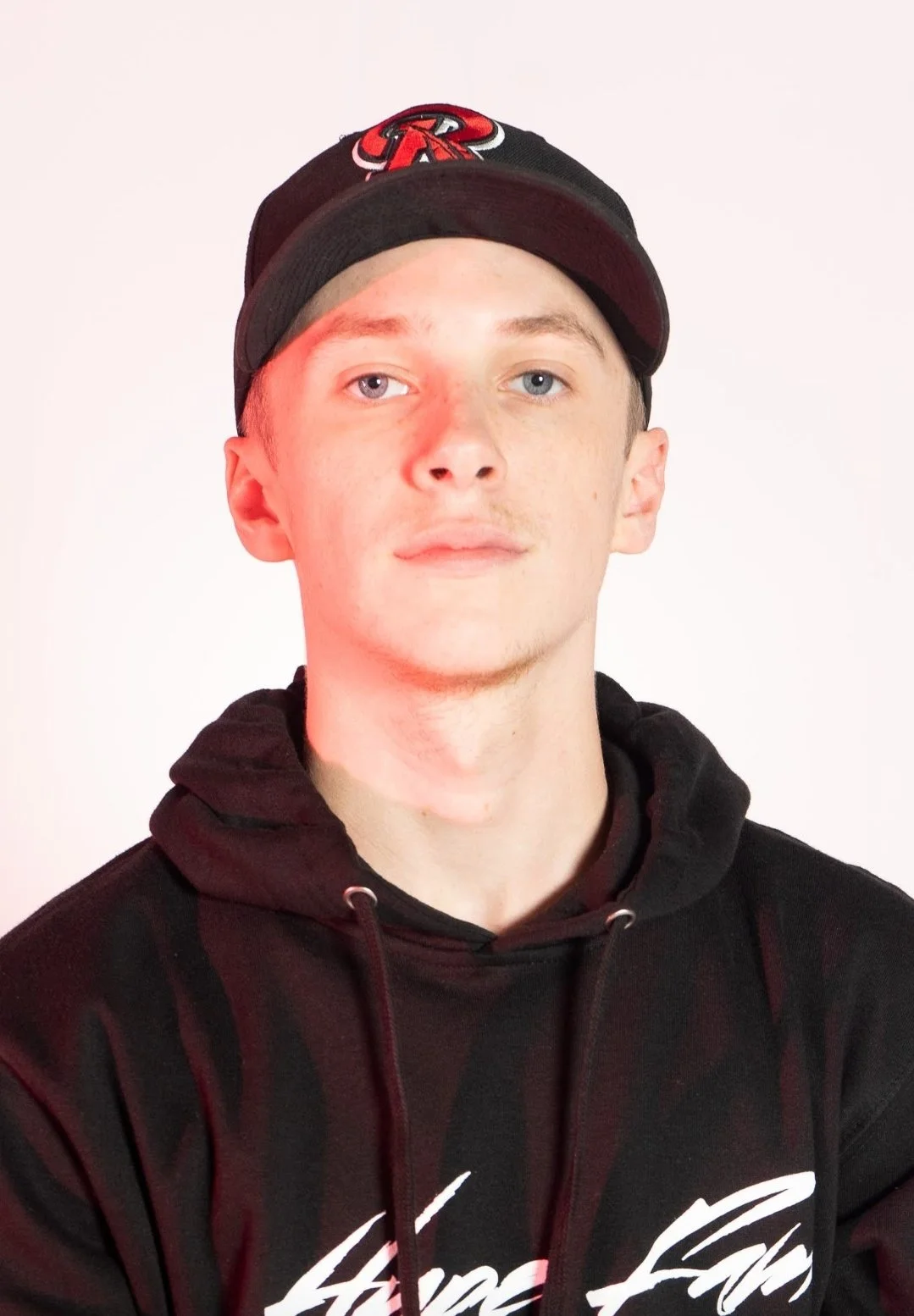 A young man with light skin, blue eyes, and short brown hair, wearing a black hoodie with white text and a black cap with a red and white logo, standing against a plain light background.