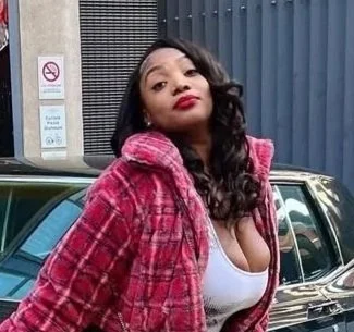 A woman with long black hair and red lipstick standing outside, wearing a pink plaid jacket and a white top, with a city street and parking lot in the background.