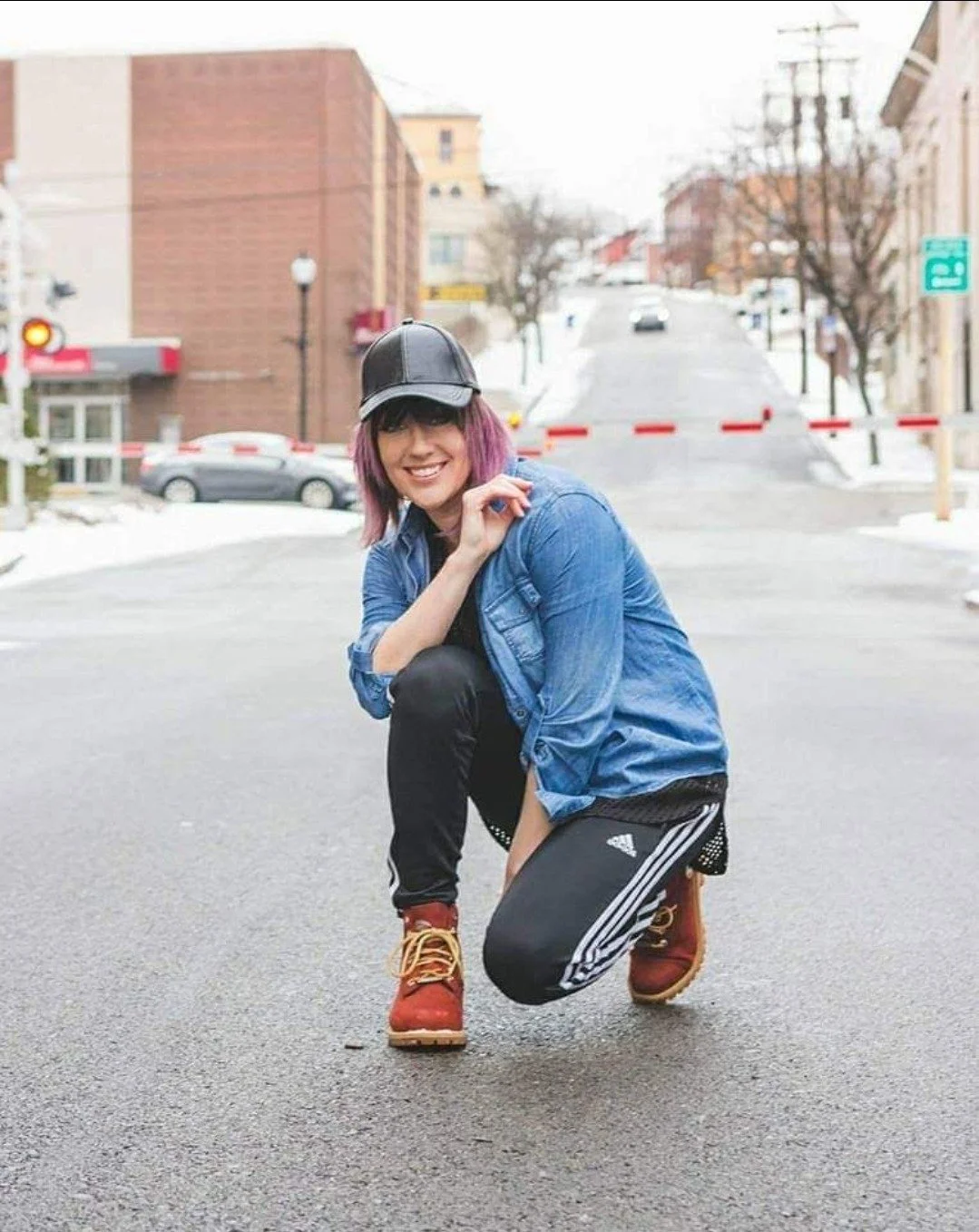 A woman with purple hair wearing a black baseball cap, blue denim jacket, black leggings with white stripes, and red boots, crouching on an urban street with parked cars and buildings in the background, smiling at the camera.