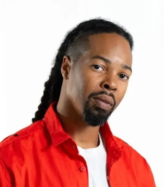 Portrait of a man with shoulder-length dreadlocks, a beard, and mustache, wearing a red button-up shirt over a white t-shirt, looking at the camera against a plain light background.