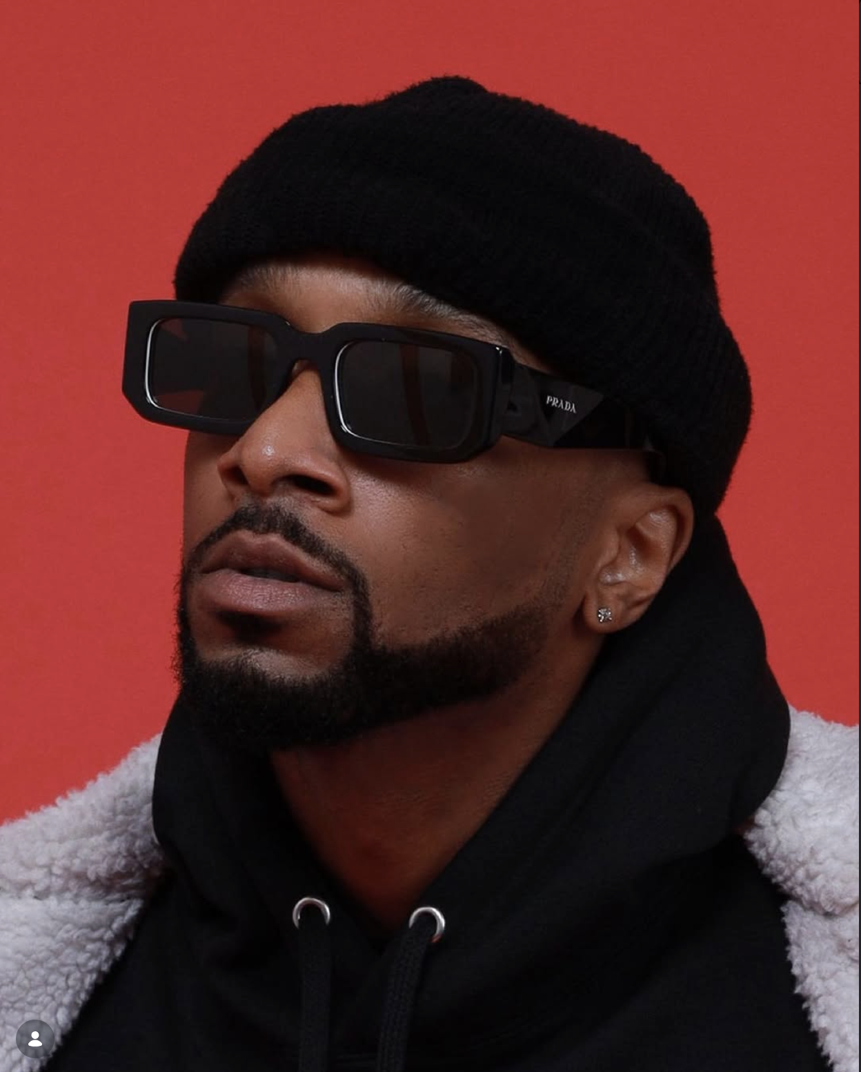 A man wearing black Prada sunglasses, a black beanie, earrings, and a hoodie with a fluffy white coat against a red background.
