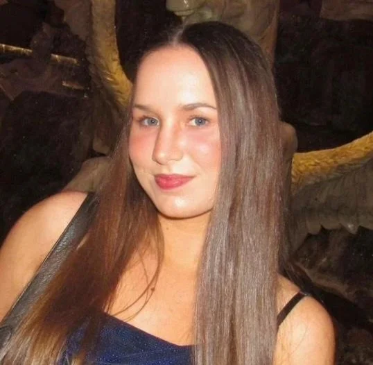 A woman with long brown hair, wearing a dark blue top and makeup, posing indoors with a smile.