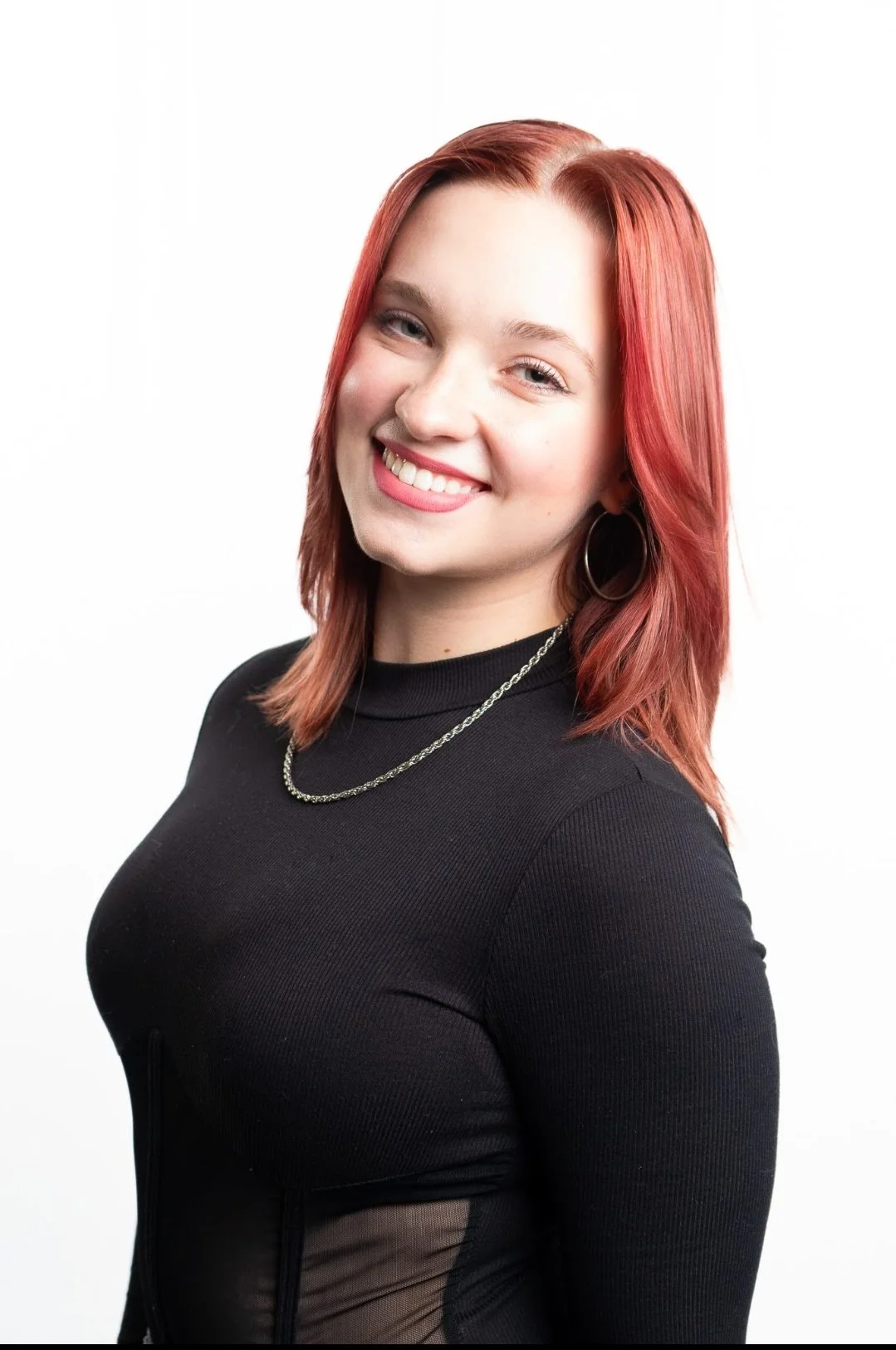 A young woman with shoulder-length red hair, smiling, wearing a black top, a necklace, and hoop earrings, against a white background.