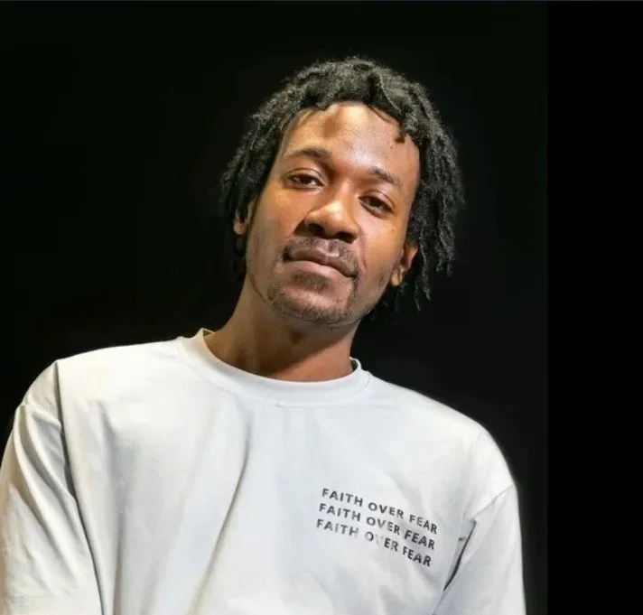 A man with dark skin and dreadlocks wearing a white t-shirt with the text "FAITH OVER FEAR" repeated three times, standing against a black background.