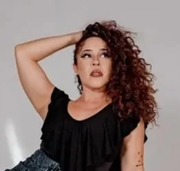 Woman with curly red hair wearing a black top, posing with one hand behind her head.
