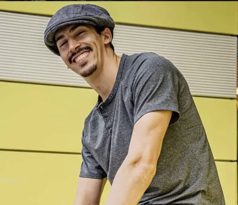 A smiling man wearing a gray T-shirt and a gray flat cap, standing in front of a yellow background with horizontal lines.