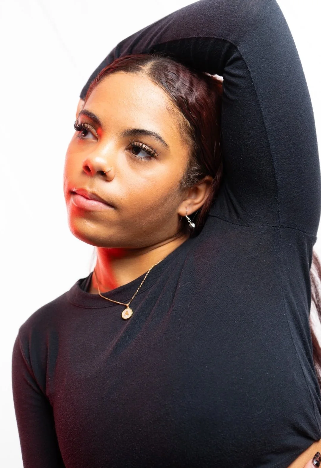 A young woman with medium brown skin and long, dark hair with reddish highlights, wearing a black long-sleeve top, a small necklace with a pendant, and earrings, posing with her right arm lifted behind her head against a white background.