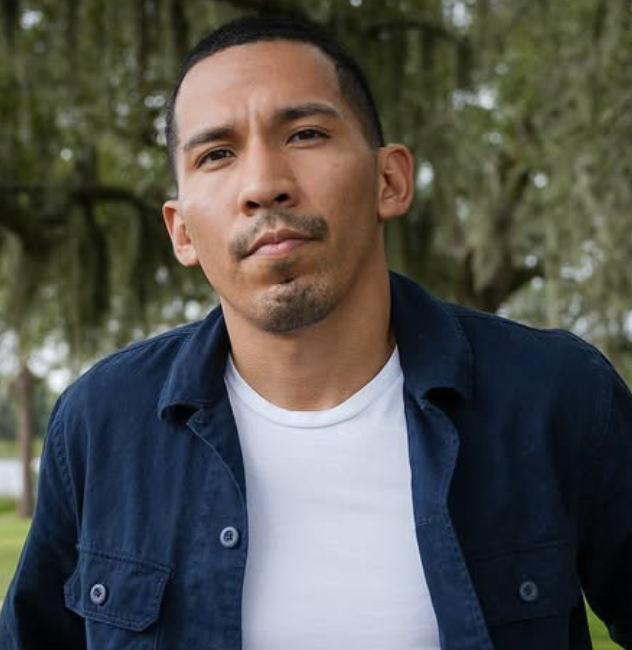 A man with short black hair, wearing a white T-shirt and a dark blue shirt, standing outdoors with trees in the background.