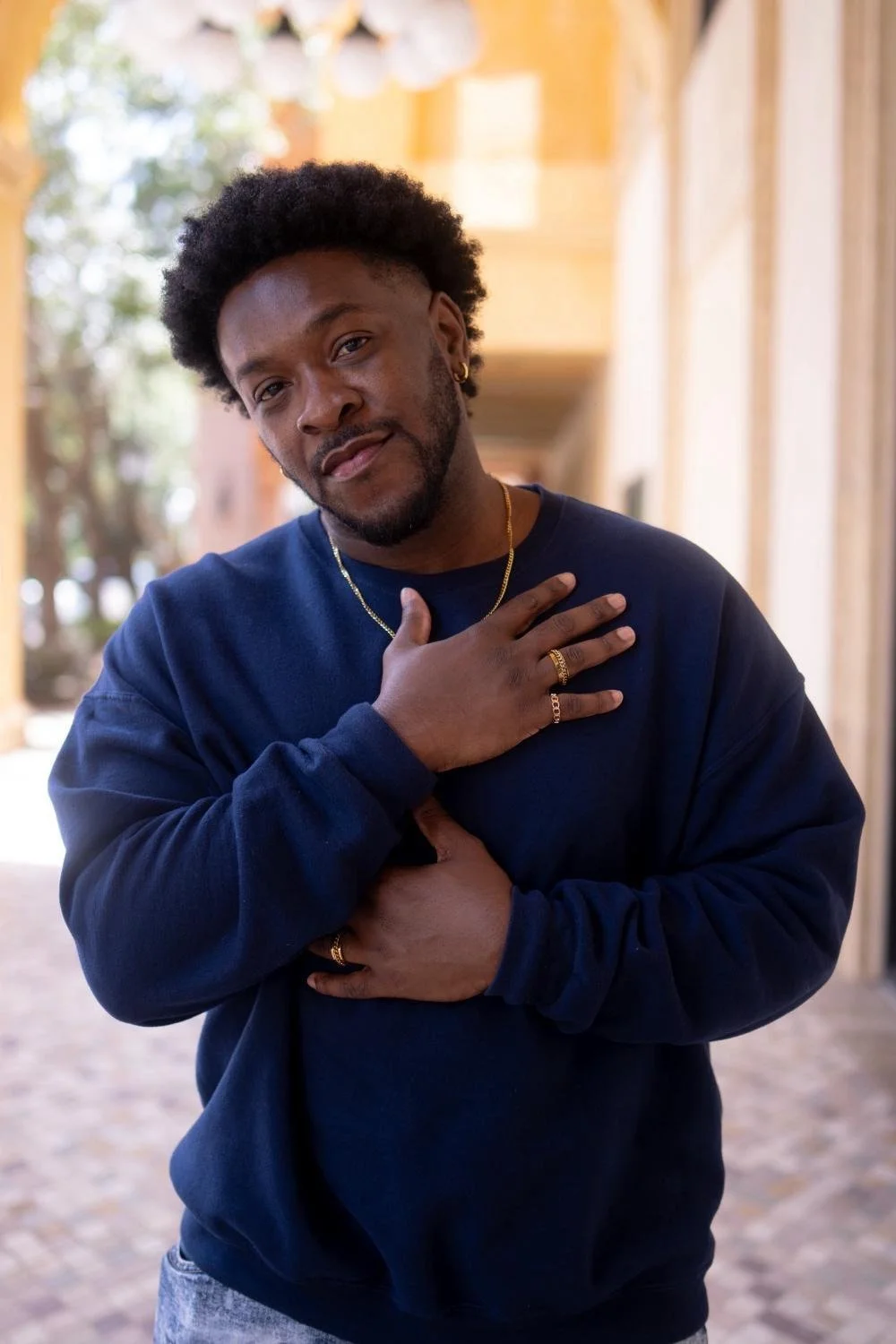 A young man with dark skin, curly hair, and a beard posing outdoors in a navy blue sweatshirt. He is touching his chest with his right hand and his stomach with his left, wearing gold rings, a gold chain, and an earring, with a slight smile. The background shows a blurred outdoor setting with trees and a building.