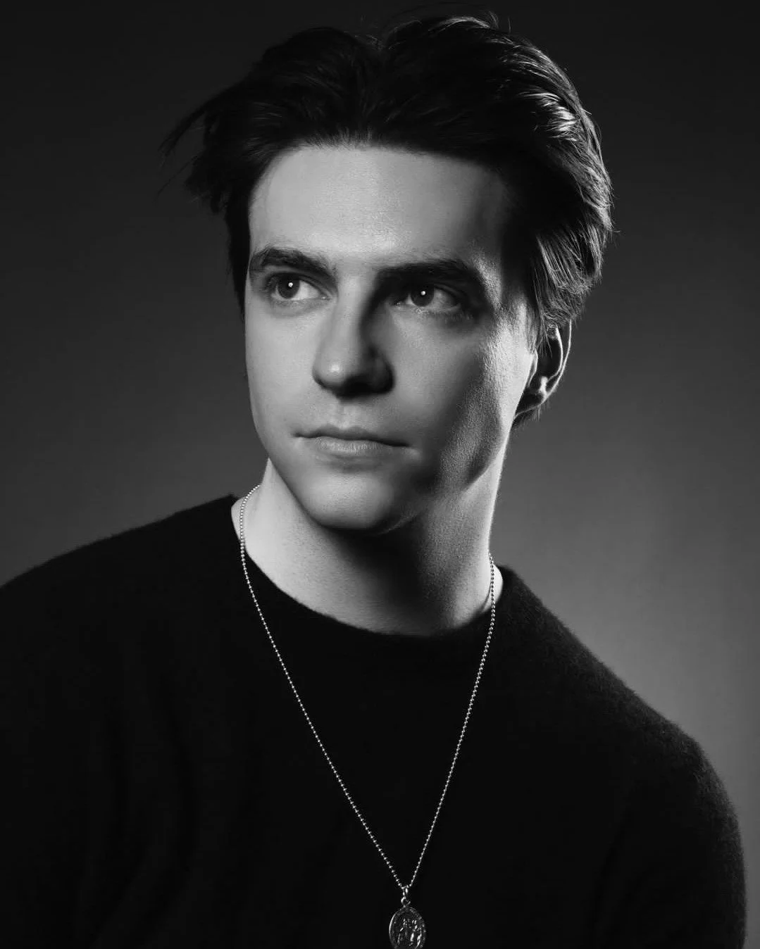 Black and white portrait of a young man with dark, tousled hair wearing a dark sweater and a pendant necklace, looking slightly to the side.
