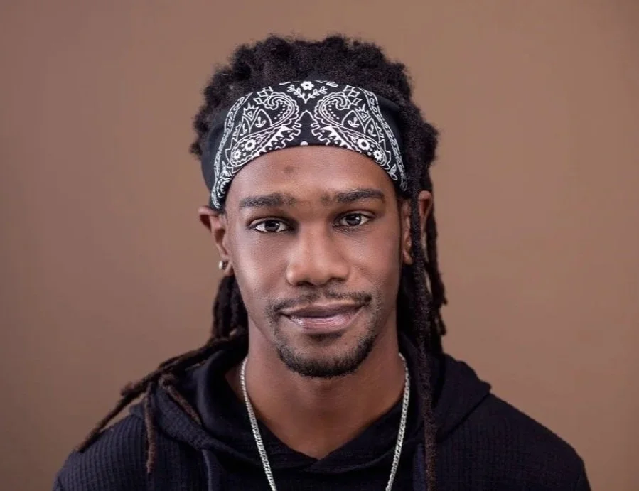 A young man with dark skin, wearing a black hoodie, a silver chain, and a black bandana with white patterns tied around his head, against a plain brown background.