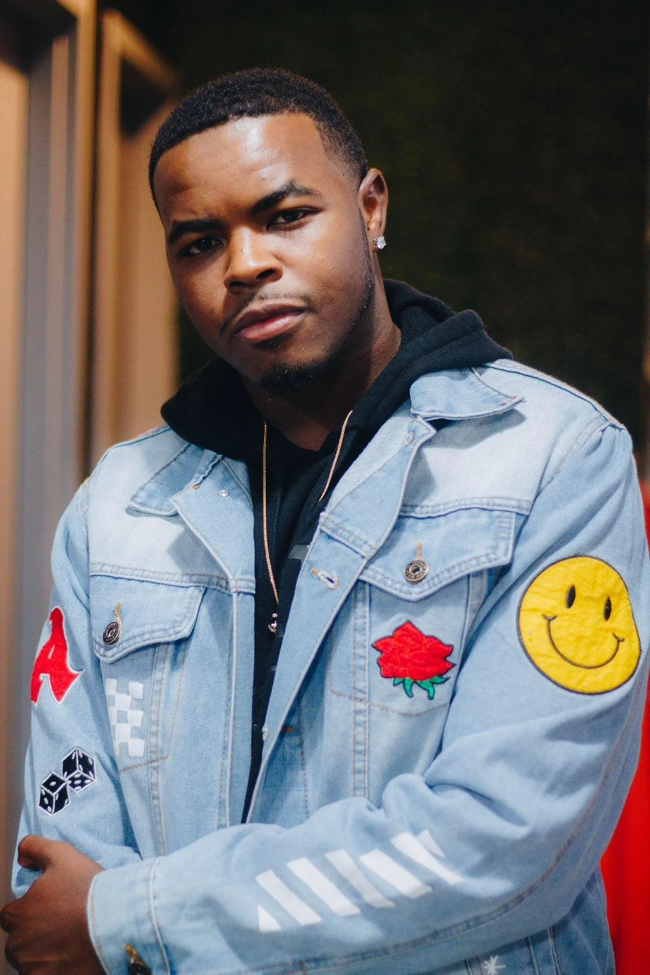 Young man with dark skin, short hair, wearing a denim jacket with colorful patches, a black hoodie, and jewelry, looking at the camera.