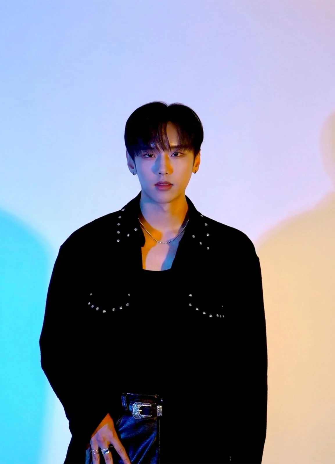 A young man with black hair wearing a black studded jacket, black shirt, and jewelry, standing against a colorful gradient background.