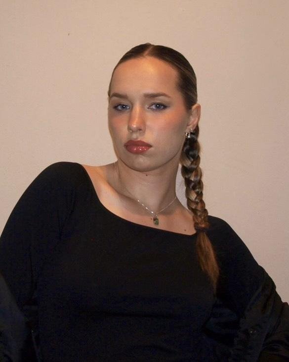 A young woman with fair skin and dark blonde hair in a braid, wearing a black top, silver necklace, and earrings, posing against a plain beige wall.