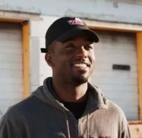 A man with dark skin wearing a black cap and a gray hoodie, smiling outdoors in front of a building.