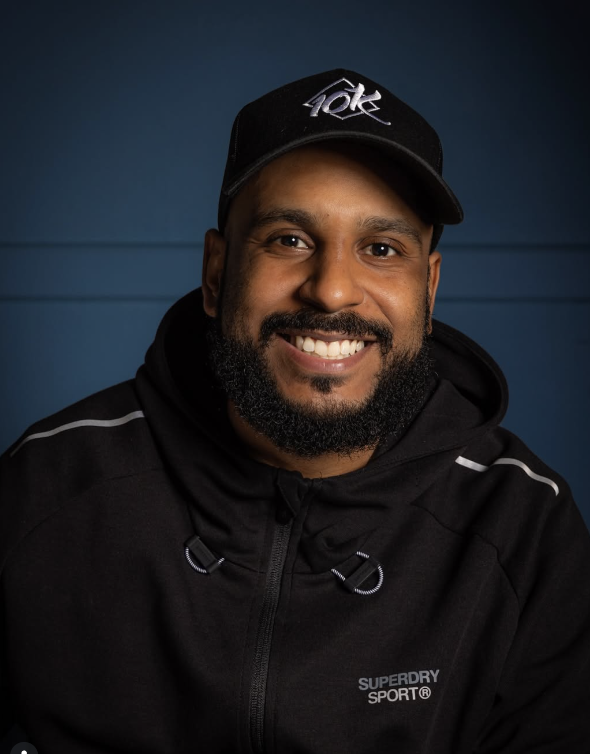 A man with a beard and mustache wearing a black cap and black jacket, smiling at the camera against a blue background.