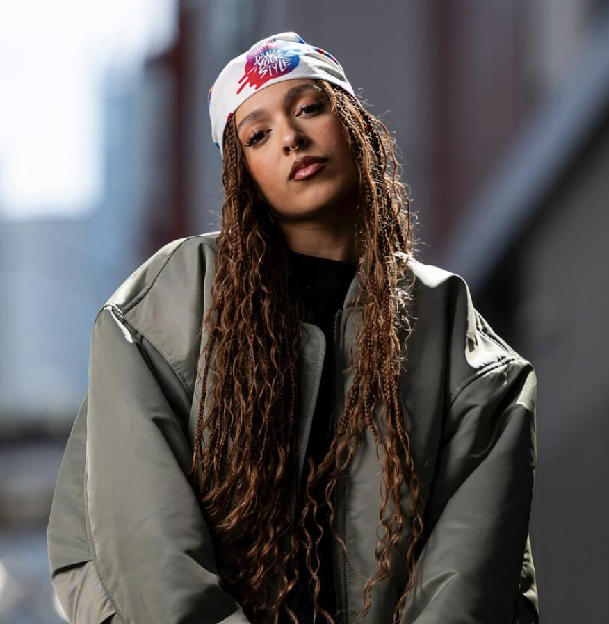 A woman with long, curly brown hair and a nose piercing, wearing a gray jacket and a white bandana with colorful graphics, standing outdoors with blurred background.