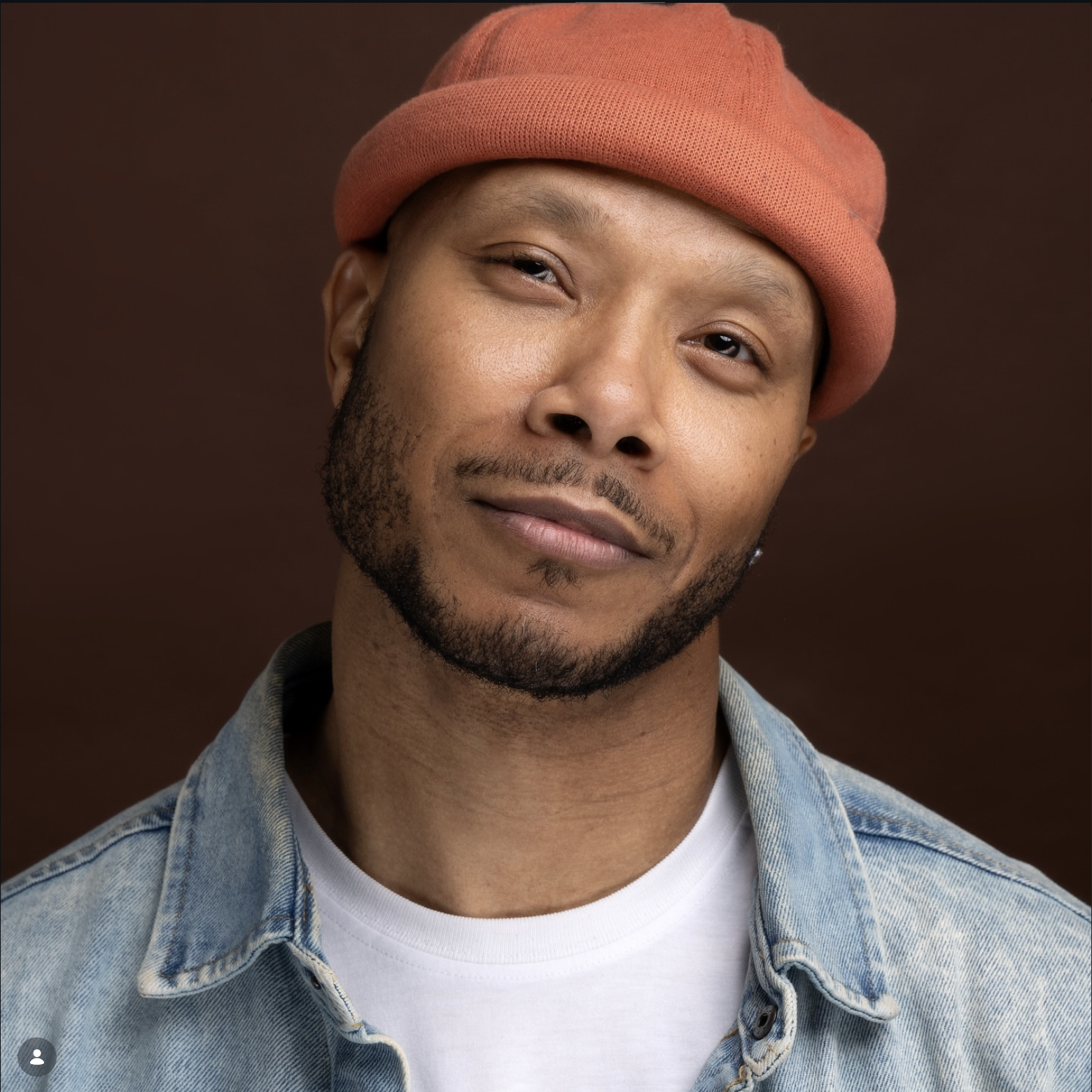 A man wearing a light denim jacket, white t-shirt, and a pink beanie, smiling softly against a plain dark background.