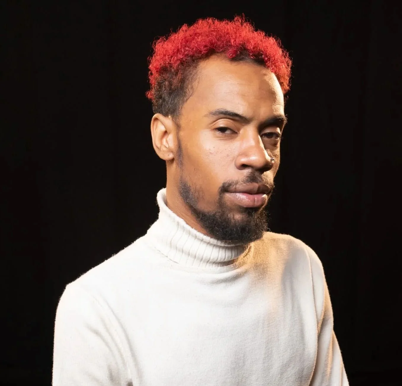 A man with red curly hair, a beard, and mustache wearing a white turtleneck sweater against a black background.